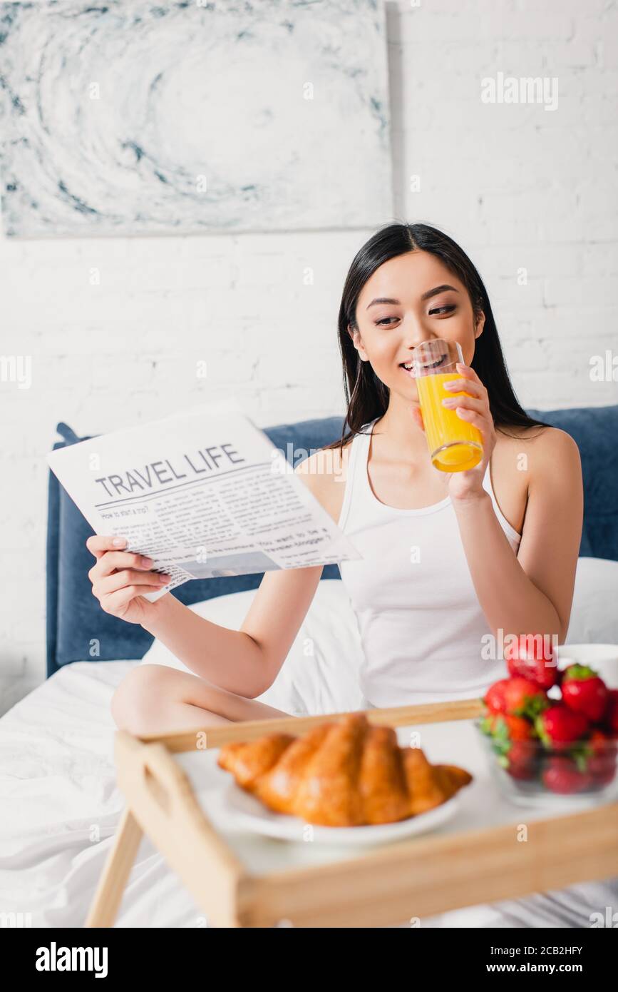 Fuoco selettivo di sorridente donna asiatica che legge giornale con il viaggio scritta life e bicchiere di succo d'arancia sul letto Foto Stock
