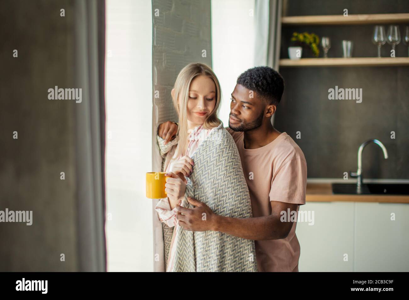 Bel ragazzo africano di baciare la sua ragazza caucasica sul collo dal retro, in piedi a casa in cucina camera con arredamento moderno Foto Stock
