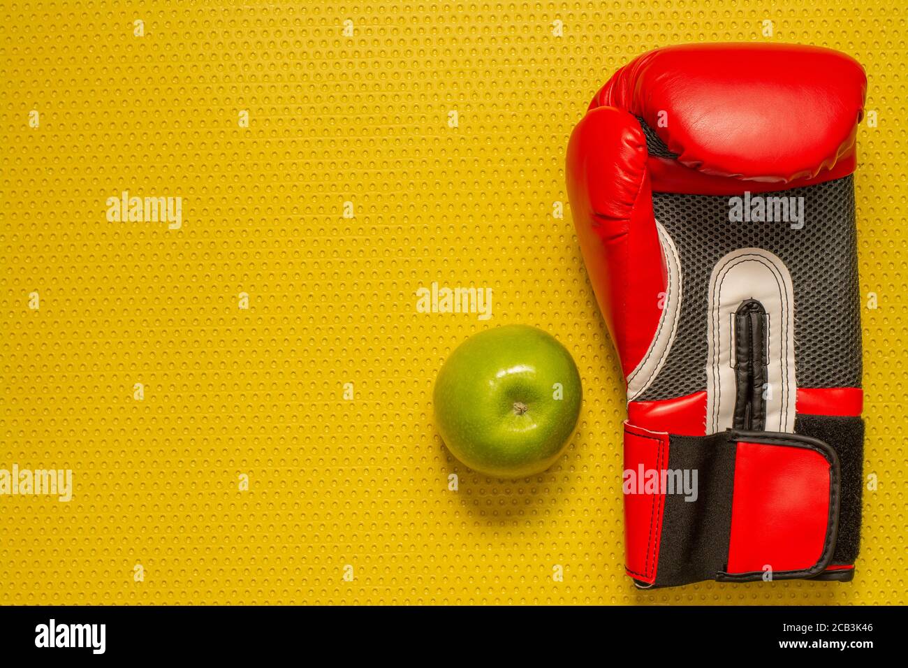 Vista dall'alto di una mela verde e di un guanto di boxe rosso sul tappetino giallo. Sport a casa, allenamento fitness Foto Stock