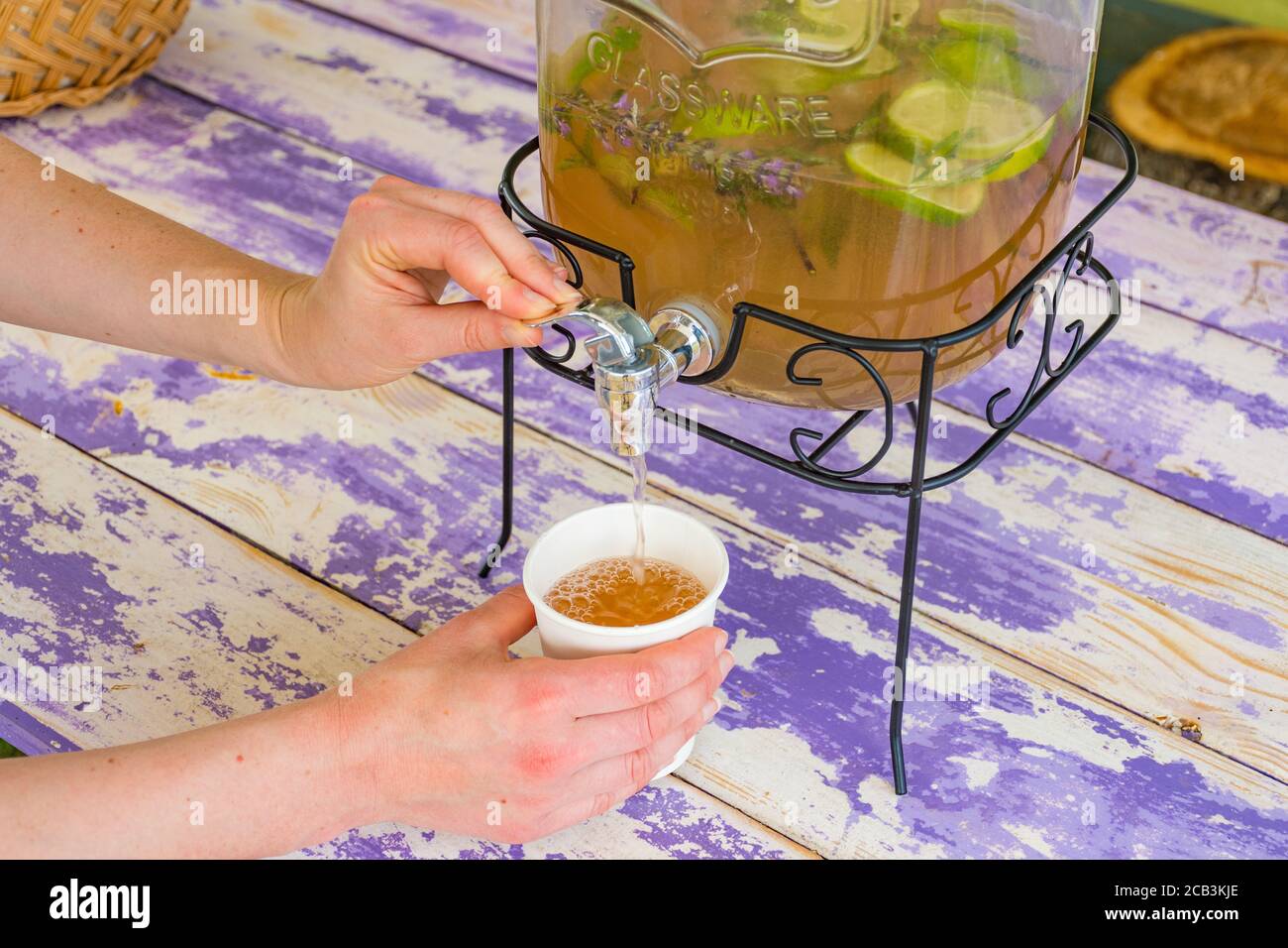 Limonata fatta in casa in vaso di vetro vintage con fette di lavanda, limoni e lime su un tavolo di legno, ragazza riempie un bicchiere con le mani Foto Stock