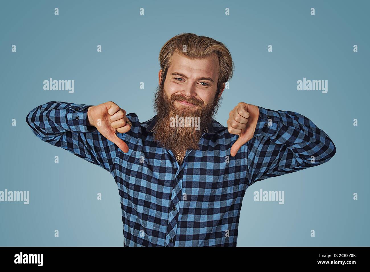 Uomo sorridente con il pollice giù. Ragazzo allegro che esprime disapprovazione. Concetto dissimile. Hipster maschio con barba in camicetta a scacchi con plaid blu che mostra il tum Foto Stock