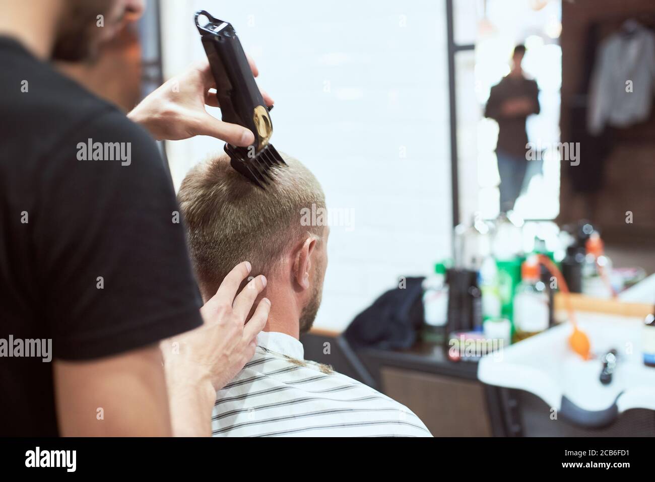 Vista ravvicinata sul retro di un uomo al barbiere che ha un taglio di capelli alla moda. Un acconciatore irriconoscibile che indossa una t-shirt nera sta lavorando con un rifinitore nero in mano Foto Stock