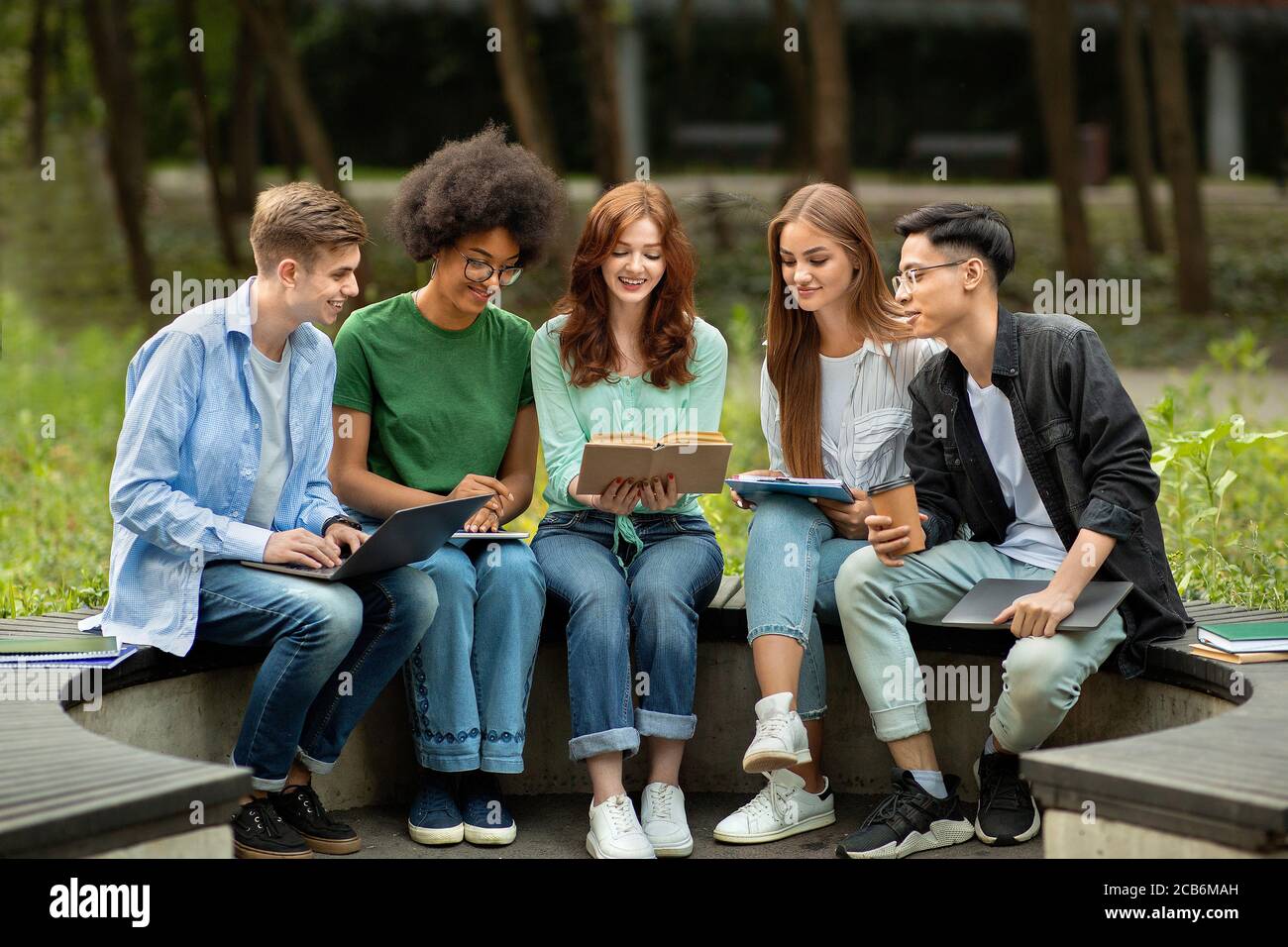 Studenti universitari multiculturali che studiano all'aperto con libri e dispositivi, preparandosi per le lezioni Foto Stock