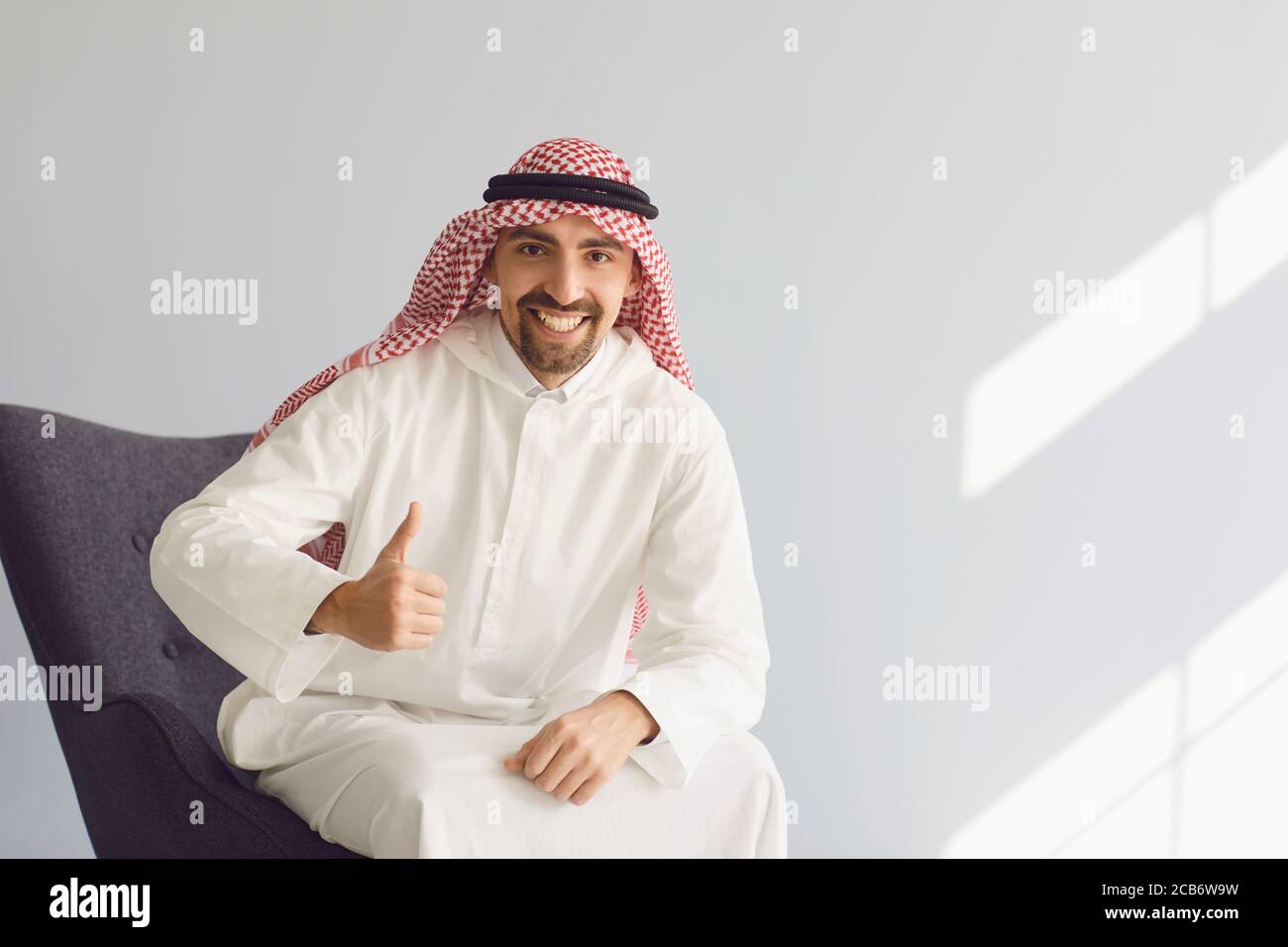 Uomo arabo sorridente mentre si siede su una sedia su uno sfondo grigio. Foto Stock