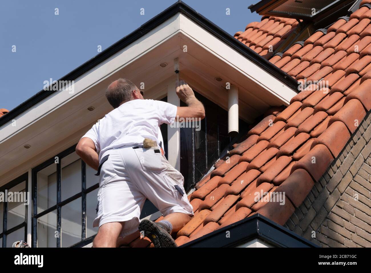 Pittore di casa professionale in piedi su una scala facendo vernice esterna di un dormer. Sfondo blu cielo. Ambiente urbano Foto Stock