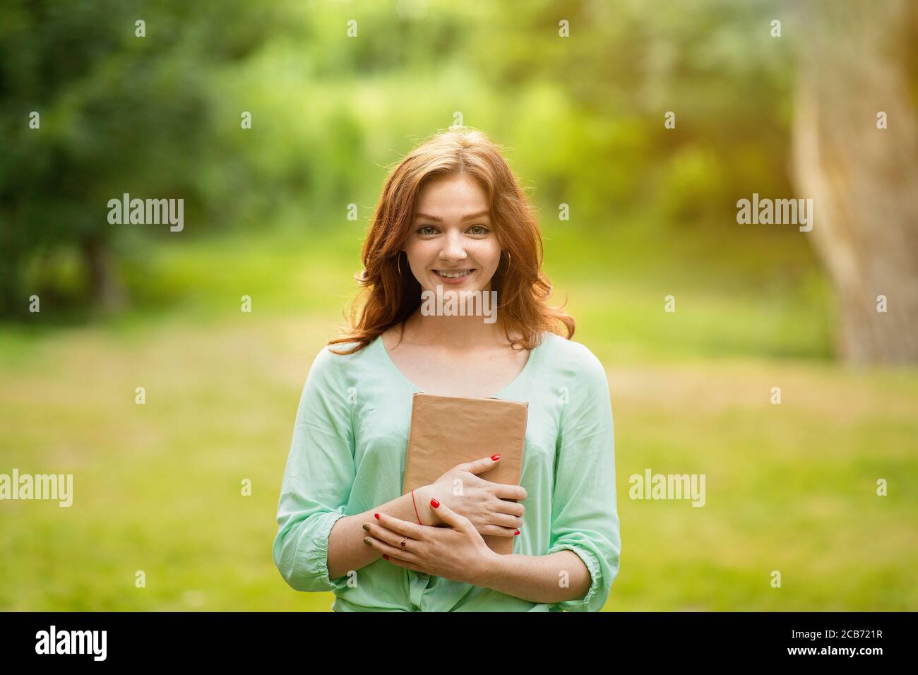 Sorridente ragazza rossa in posa con il libro in mani, godendo di leggere all'aperto Foto Stock