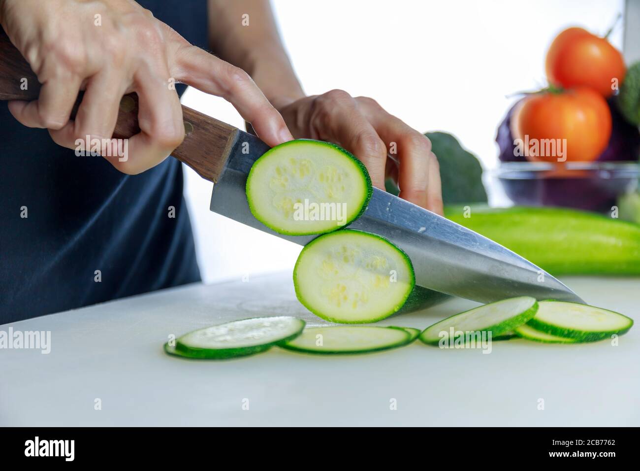 Primo piano mani donne che tengono coltello e taglio Courgette fresco sul tagliere. La mano femminile affetta il cetriolo giapponese grezzo in cerchi con coltello su cu Foto Stock