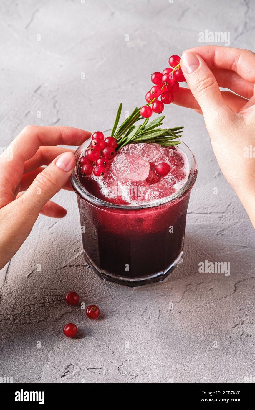 Le mani della donna contengono bacche di ribes per la preparazione di cocktail di frutta fresca ghiacciata in vetro con foglia di rosmarino, fondo di pietra in cemento Foto Stock