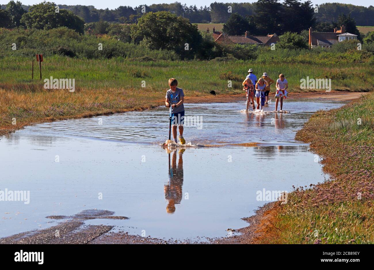 I bambini cavalcano i loro scooter attraverso l'accesso stradale allagato con la marea alluvionale al porto sulla costa nord del Norfolk a Thornham, Norfolk, Inghilterra, Regno Unito. Foto Stock