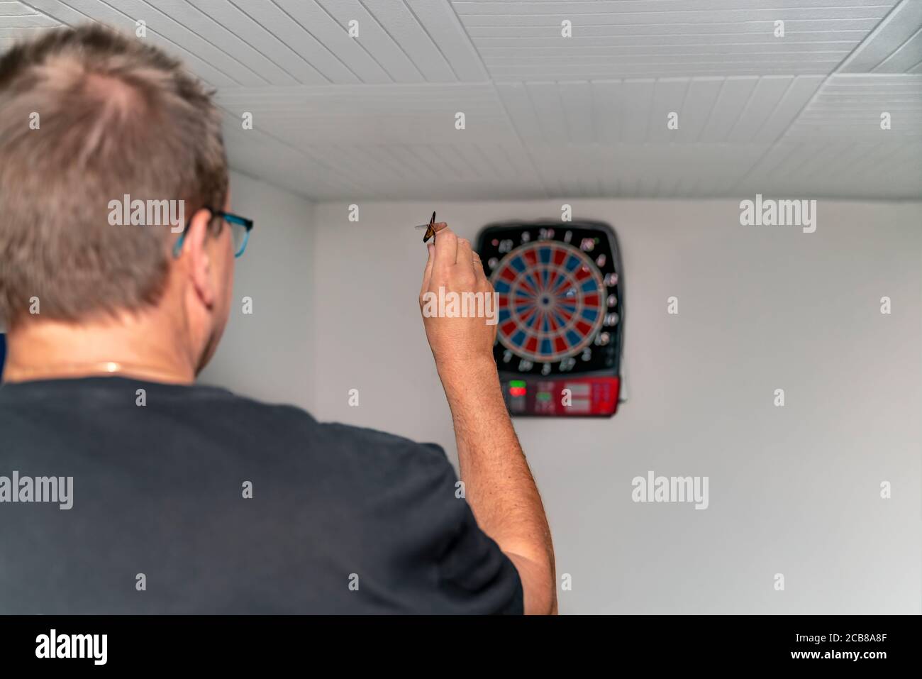 Un uomo biondo di 50 anni gioca freccette. Una macchina a freccette elettrica mostra i suoi colpi in un duello. Il dartboard è appeso in una cantina di festa. Foto Stock