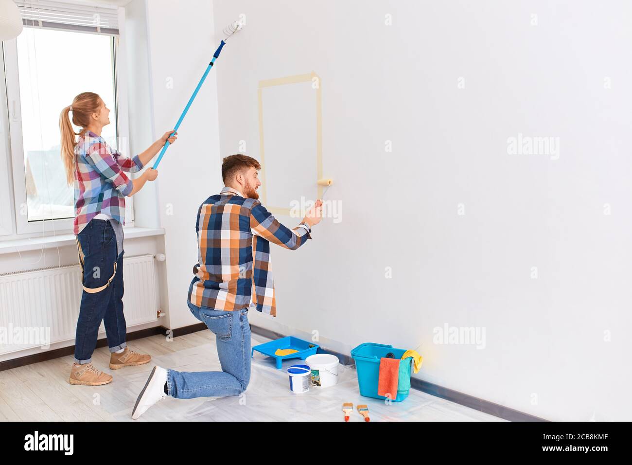 Squadra di due giovani pittori professionisti maschili e femminili in abbigliamento a scacchi con spazzole e rulli che lavorano all'interno, facendo il restauro del colore delle pareti di t Foto Stock