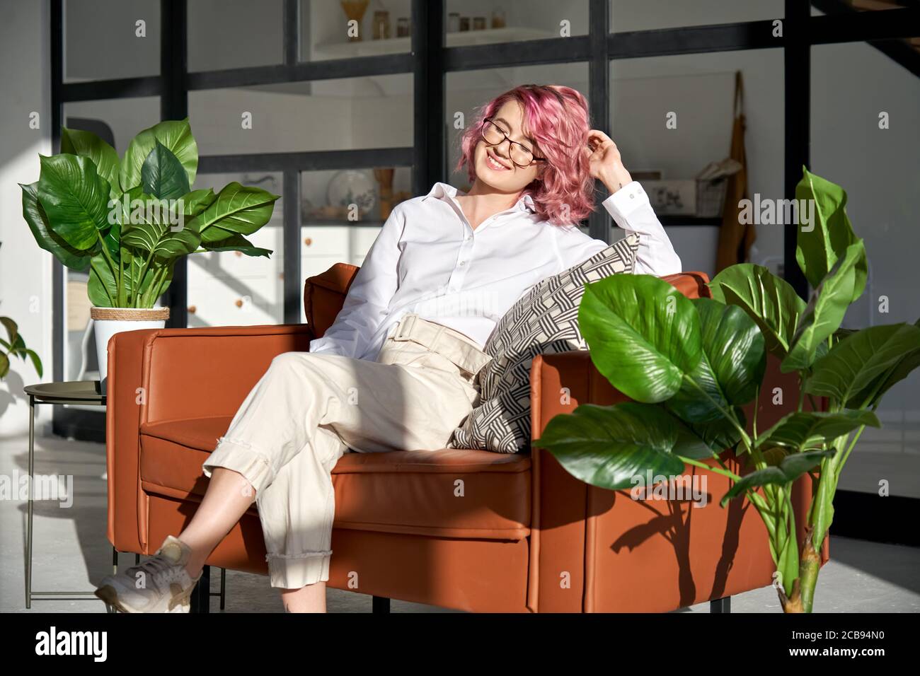 Ragazza hipster sorridente con i capelli rosa che si rilassa sul divano in un accogliente soggiorno soleggiato. Foto Stock