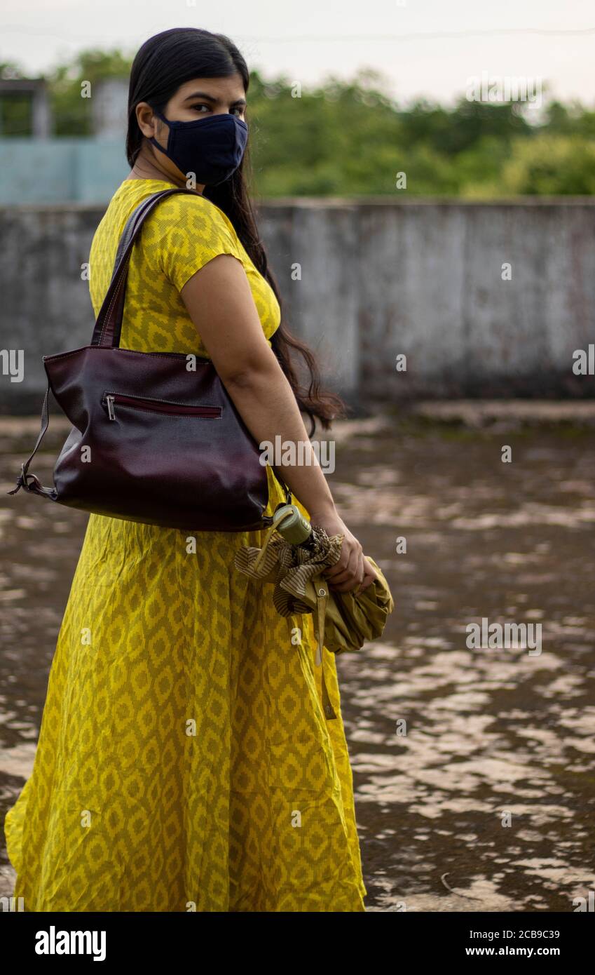 Una donna indiana in abito giallo e maschera sul viso sta andando fuori con ombrello e borsa dopo sbloccare 1.0 di pandemia del virus della corona Foto Stock