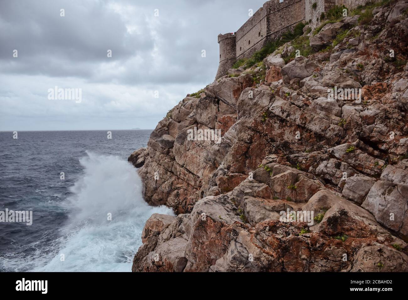 Onda che si schiantano contro le rocce con le pareti di Dubrovnik sullo sfondo, Croazia Foto Stock