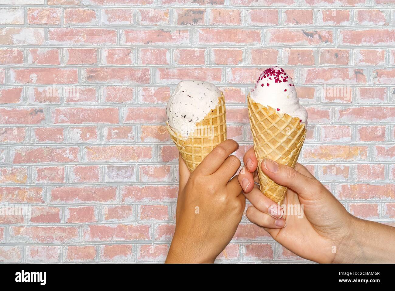 Le mani di madre e figlia tiene il mais del gelato con il gelato del latte che asseive il vecchio muro di mattone rosso. Due mani tiene i gelati. Foto Stock