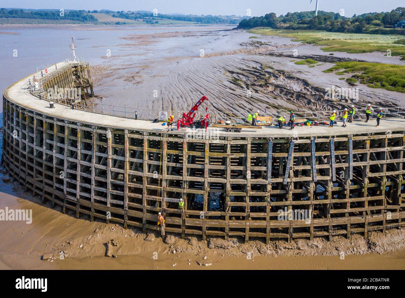 Lavoratori del canale e del fiume Trust in discesa a bassa marea per effettuare riparazioni al molo di legno all'ingresso di Sharpness New Dock sul fiume Severn in Gloucestershire. Canali di beneficenza Canal e River Trust stanno utilizzando una gru a ragno per portare in tavole di quercia greenheart che saranno fissate in posizione da una squadra abbassata su corde sospese sopra il fangoso letto del fiume a bassa marea. Foto Stock