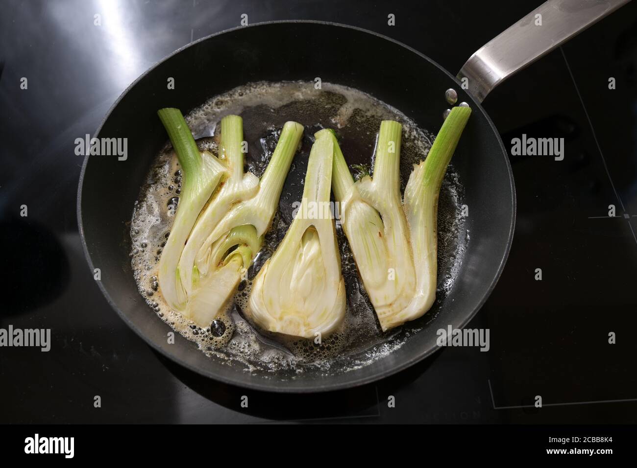 Le fette di finocchio fresco vengono fritte nel burro in una padella di cottura su un fornello nero, preparazione per un pasto vegetariano sano, fuoco selezionato Foto Stock
