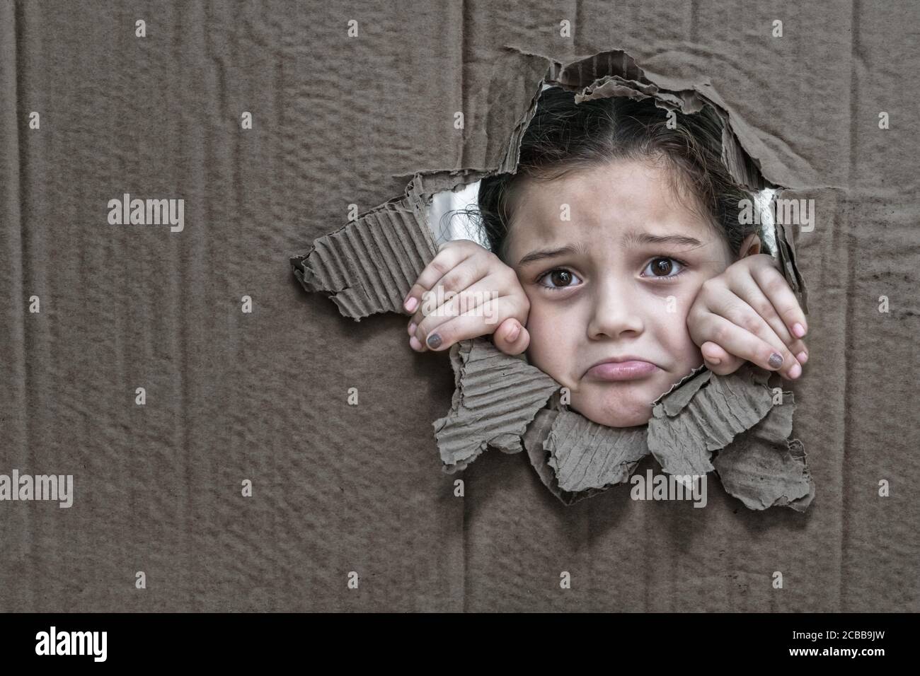 Bambina con un'espressione triste dietro un cartone, guardando attraverso un grande buco nel cartone con le sue mani che stringono il bordo del foro Foto Stock