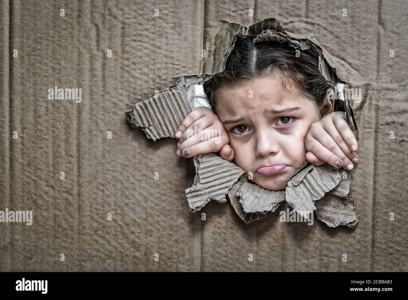 Bambina con un'espressione triste dietro un cartone, guardando attraverso un grande buco nel cartone con le sue mani che stringono il bordo del foro Foto Stock