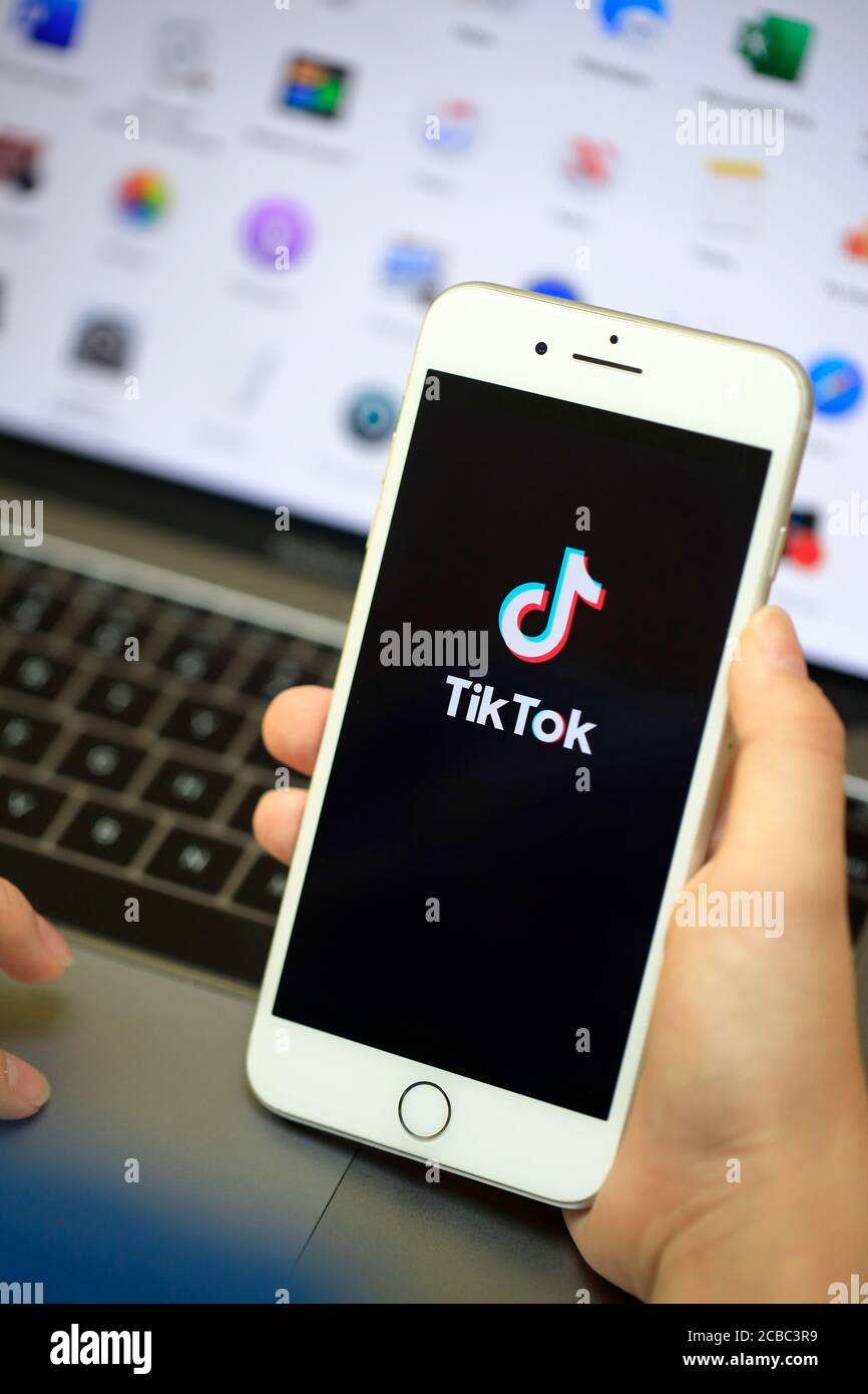 Una mano che tiene un iPhone con il logo di online App di condivisione video e hosting Tik Tok sullo schermo davanti a un computer Foto Stock