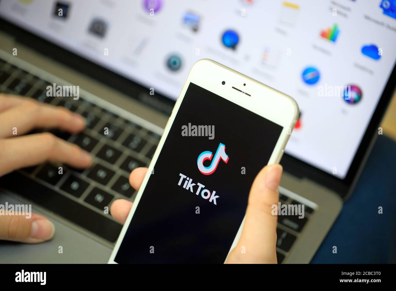Una mano che tiene un iPhone con il logo di online App di condivisione video e hosting Tik Tok sullo schermo davanti a un computer Foto Stock