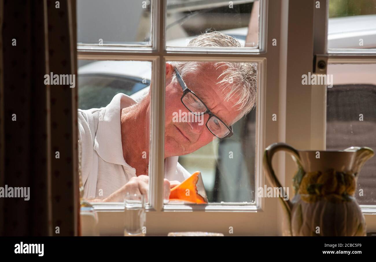 Hampshire, Inghilterra, Regno Unito. 2020. Pittore utilizzando un raschietto per preparare le finestre per la pittura vista dall'interno della proprietà. Foto Stock