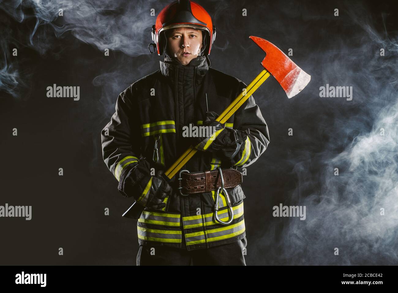 ritratto di serio e sicuro pompiere caucasico regge martello, indossando speciale uniforme protettiva sullo sfondo fumoso Foto Stock