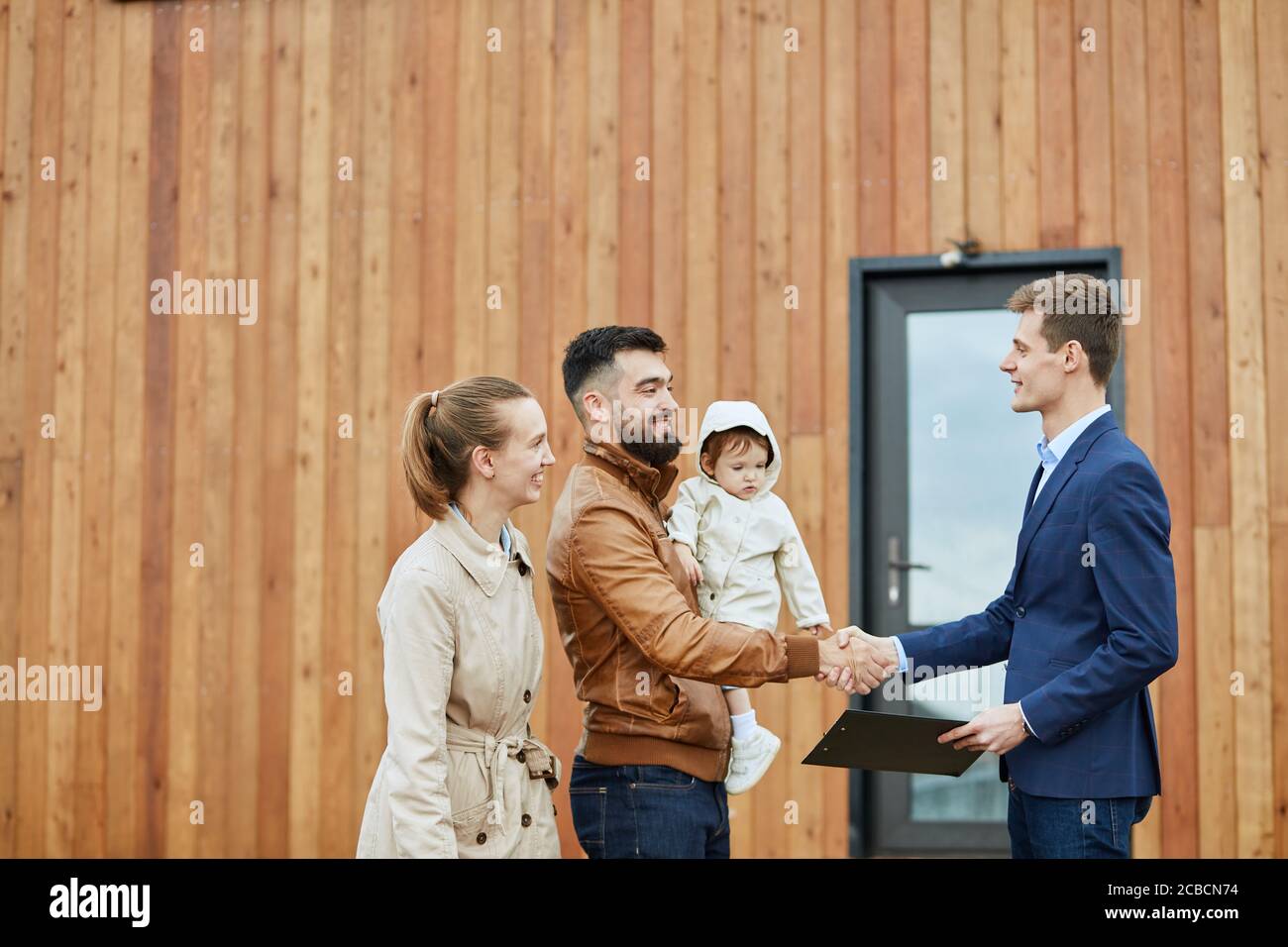 Famiglia caucasica felice e realtor indossare abito blu stand all'aperto vicino a casa nuova appena venduto. Gli uomini scuotono le mani. Foto Stock