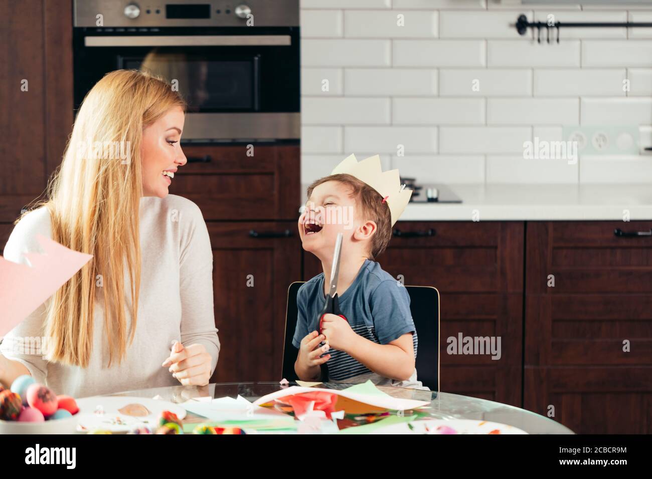 Felice giovane madre che passa il tempo con il suo gioioso figlio, seduto al tavolo da cucina con un sacco di carta colorata e uova dipinte. I preparativi di Pasqua sono io Foto Stock