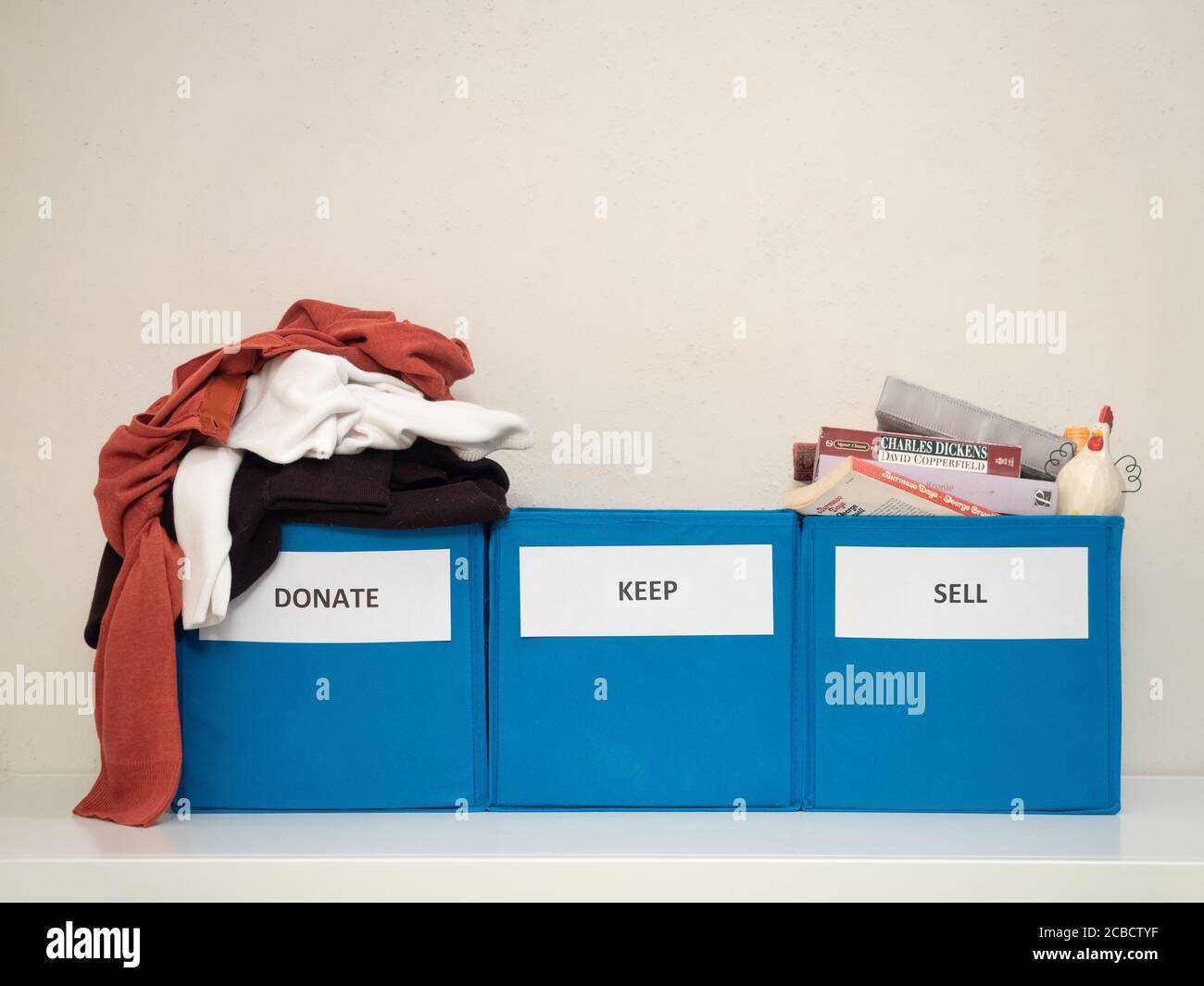 Tre bidoni turchesi etichettati donare, vendere e tenere contro una parete bianca che hanno scartato vestiti, libri classici, e un gallo. Contenitori utilizzati per dow Foto Stock