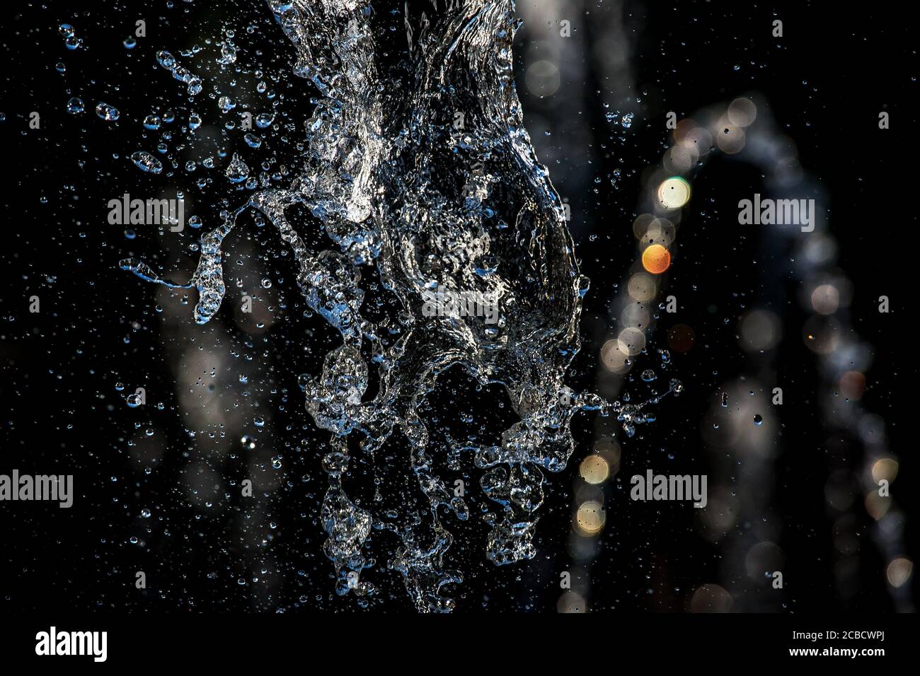Schizzi di liquidi e gocce d'acqua volanti sotto la luce del sole su sfondo nero. Foto Stock