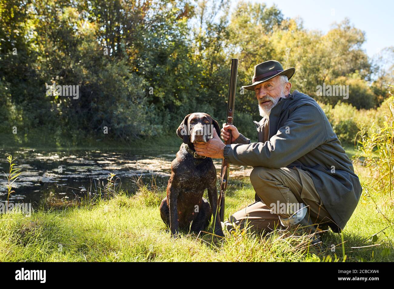 Uomo anziano dall'aspetto bello soddisfatto della caccia sul lago, siediti accanto al suo cane, in foresta, giorno di sole Foto Stock