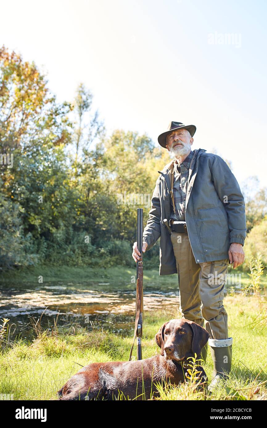 Uomo anziano di buon aspetto soddisfatto di caccia sul lago, stare accanto al suo cane, in foresta, giorno di sole Foto Stock