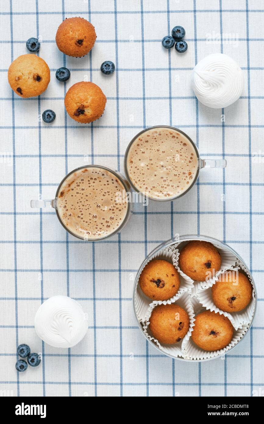 Due tazze di latte schiumoso e muffin, mirtilli, marshmallows su una tovaglia a scacchi. Concetto di colazione. Design piatto Foto Stock
