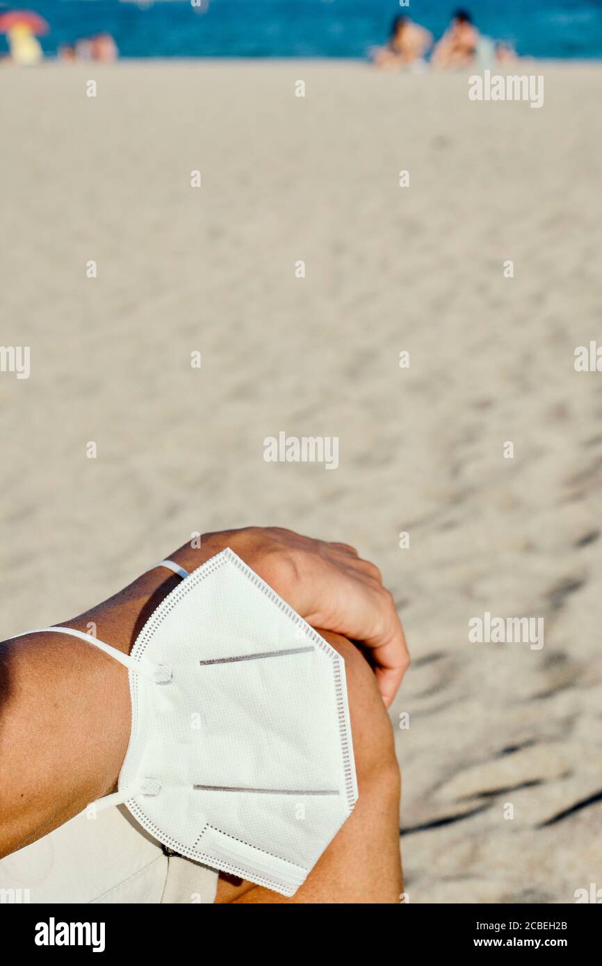 primo piano di un giovane caucasico, con calzoncini corti, seduto sulla spiaggia, con una maschera bianca al polso, come lo fa un respiratore di indossarla, Foto Stock