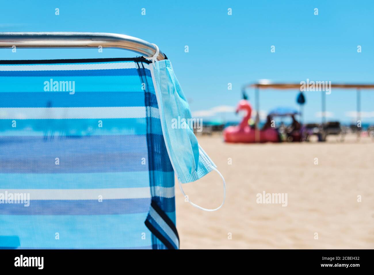 primo piano di una maschera chirurgica appesa ad una sdraio a strisce blu sulla spiaggia, con alcuni ombrelloni e il mare sullo sfondo Foto Stock