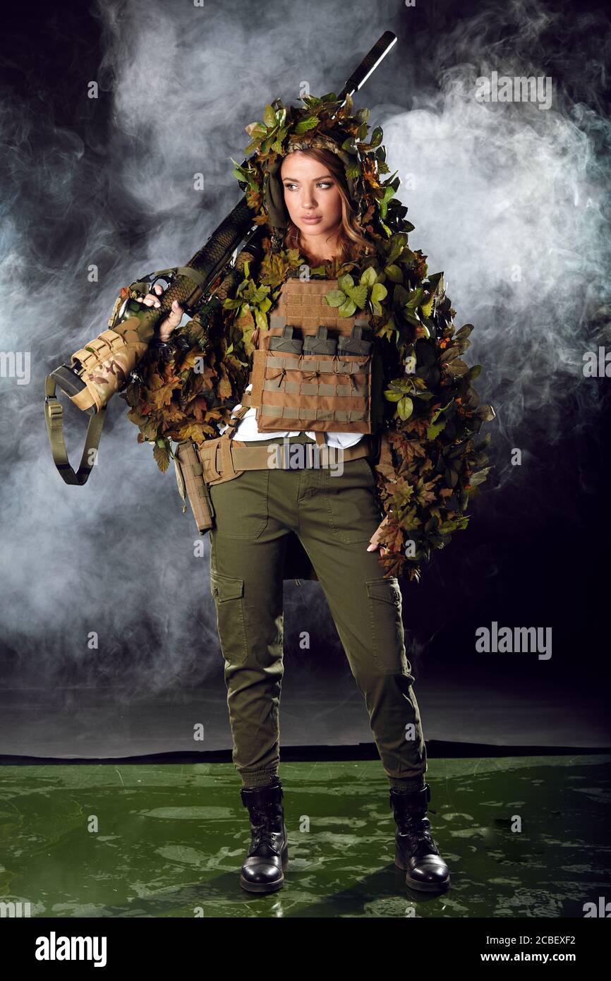 Cecchino donna camuffato in tuta ghillia in posa con fucile in notte nebbia. Armi speciali e concetto Donna. Foto Stock