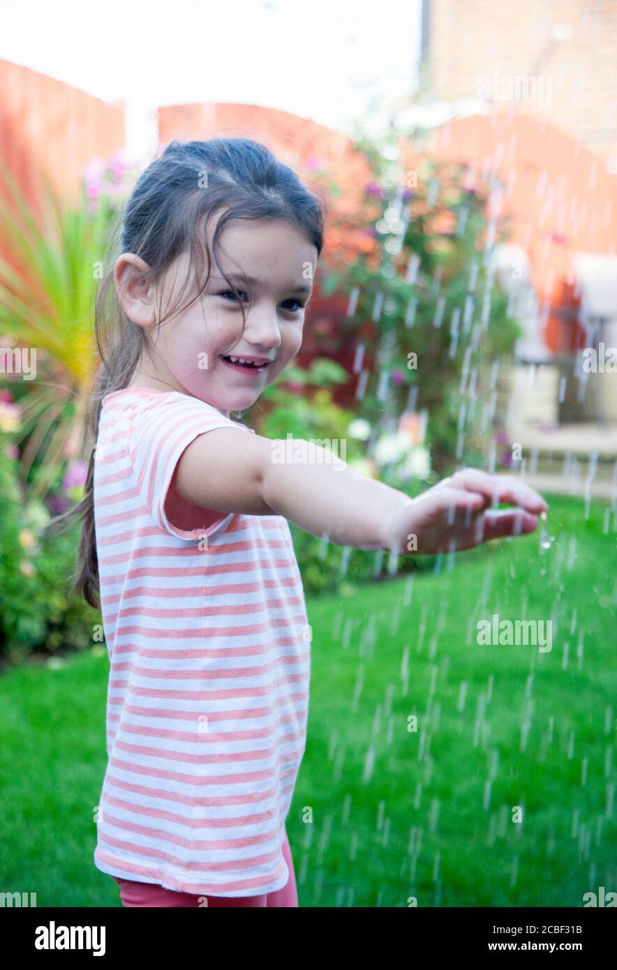 La ragazza di 4 anni, indossando una t-shirt stripea, tiene un braccio sotto le gocce d'acqua in un giardino inglese durante l'estate. Lancashire, Inghilterra, Regno Unito Foto Stock