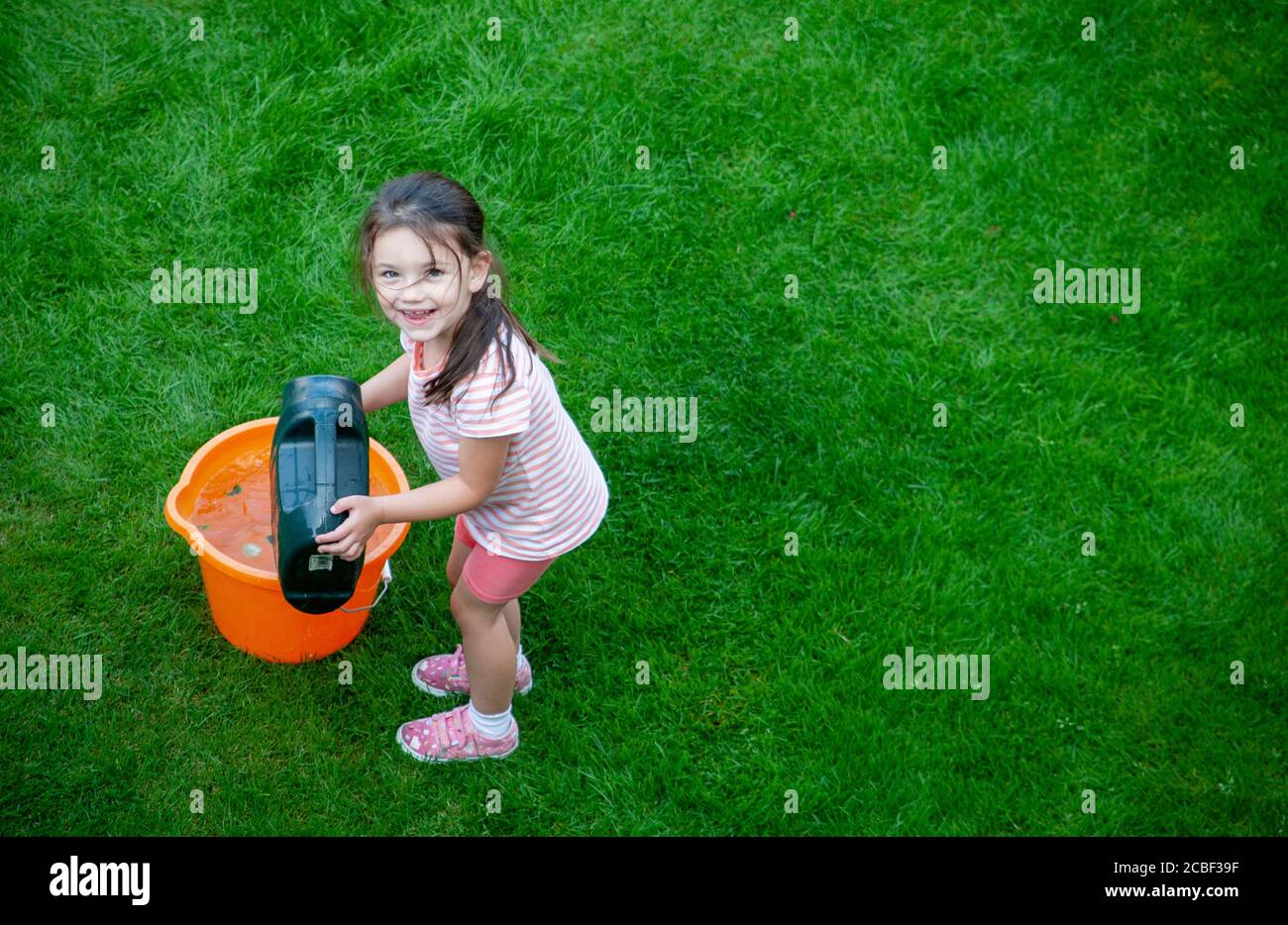 Bambina di 4 anni, indossando abiti estivi, versando acqua da una lattina di annaffiatura verde in un secchio arancione in un giardino inglese. Lancashire Regno Unito Foto Stock