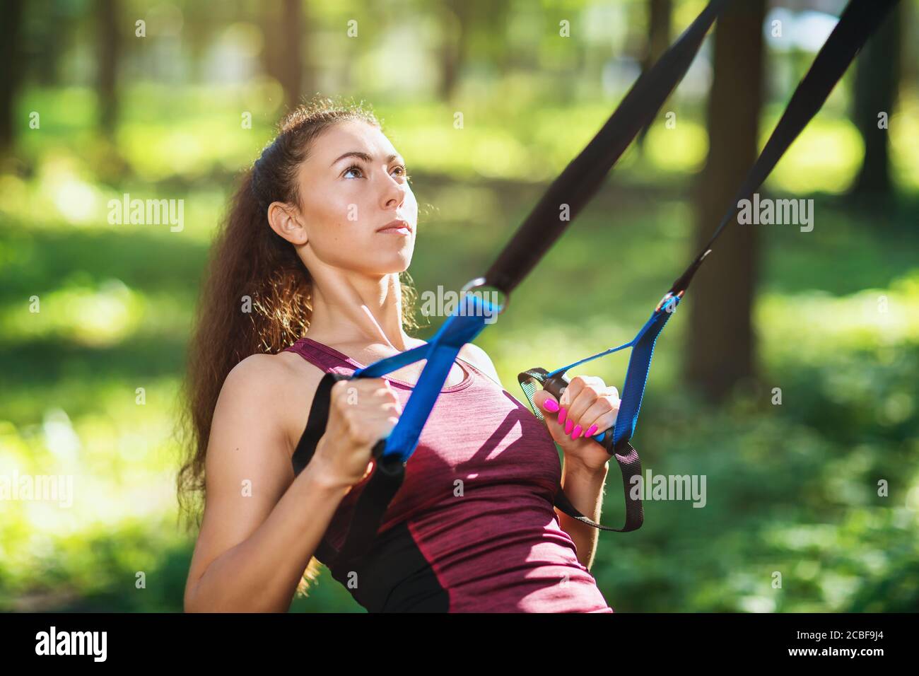 Un'attraente ragazza dai capelli marroni con i capelli lunghi in una tuta sta facendo esercizi nel parco usando un addestratore di sospensione. Atleta allena il suo muscl posteriore Foto Stock