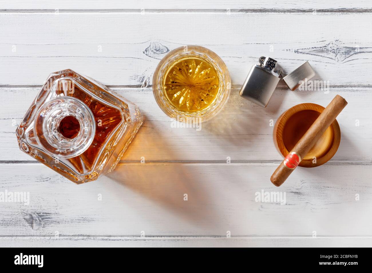 Vista di un vetro di cristallo e di un decanter pieno di whisky dorato, sigaro e accendisigari a benzina, sparati dall'alto su uno sfondo di legno bianco in difficoltà Foto Stock