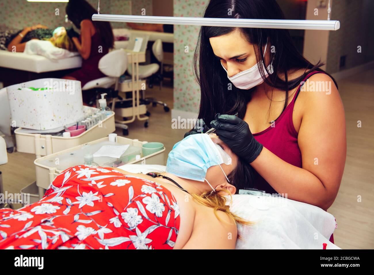 Giovane donna bruna dai capelli lunghi che mette le ciglia false sulla signora in salone di bellezza con maschere, nuovo normale Foto Stock