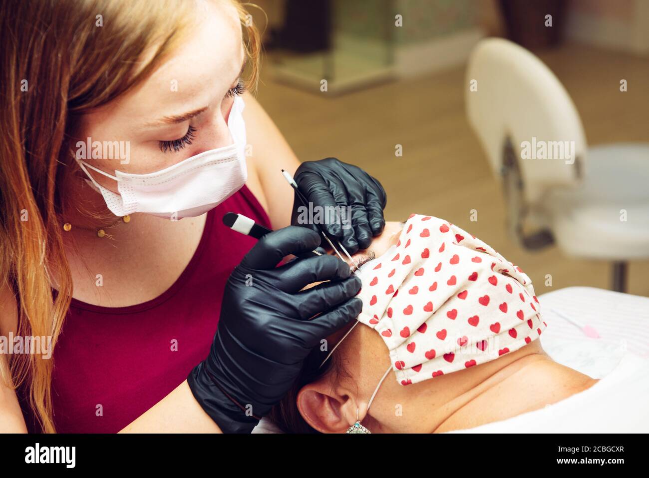Giovane donna bionda dai capelli lunghi che mette sulle ciglia false a signora in salone di bellezza con maschere, nuovo normale Foto Stock