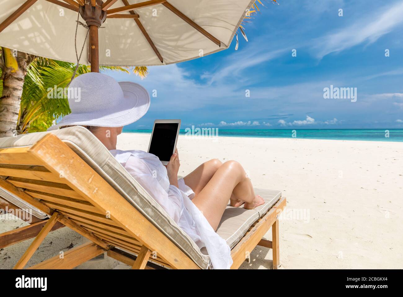 Donna con computer tablet sulla spiaggia seduta nella sdraio. Cappello giovane donna con PC tablet in spiaggia, vacanza estiva di lusso, tempo libero Foto Stock