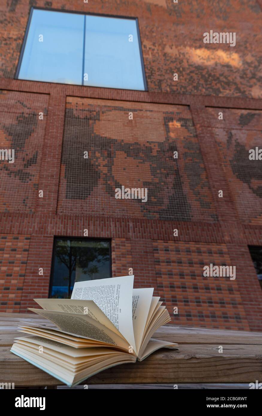 Un libro aperto poggiato su una panchina di fronte un edificio in mattoni rossi con una finestra in vetro colorato che si riflette il cielo blu Foto Stock