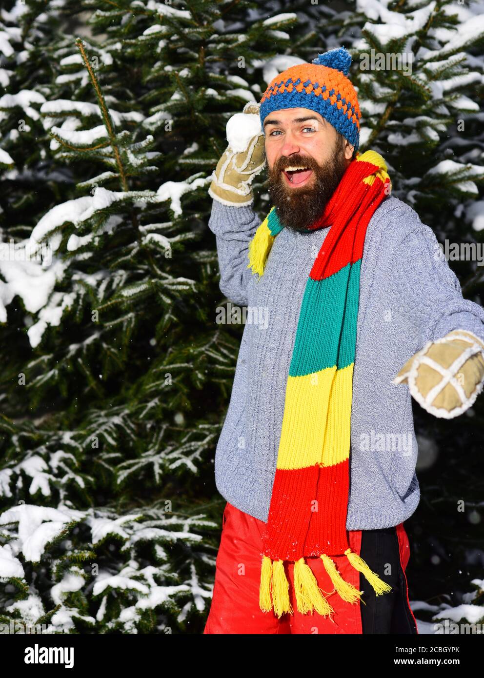 Uomo con cappello, sciarpa e guanti. Ragazzo con faccia felice con i  firtrees coperti di neve sullo sfondo. Concetto di intrattenimento  all'aperto. Macho con barba e baffi gioca con la palla di