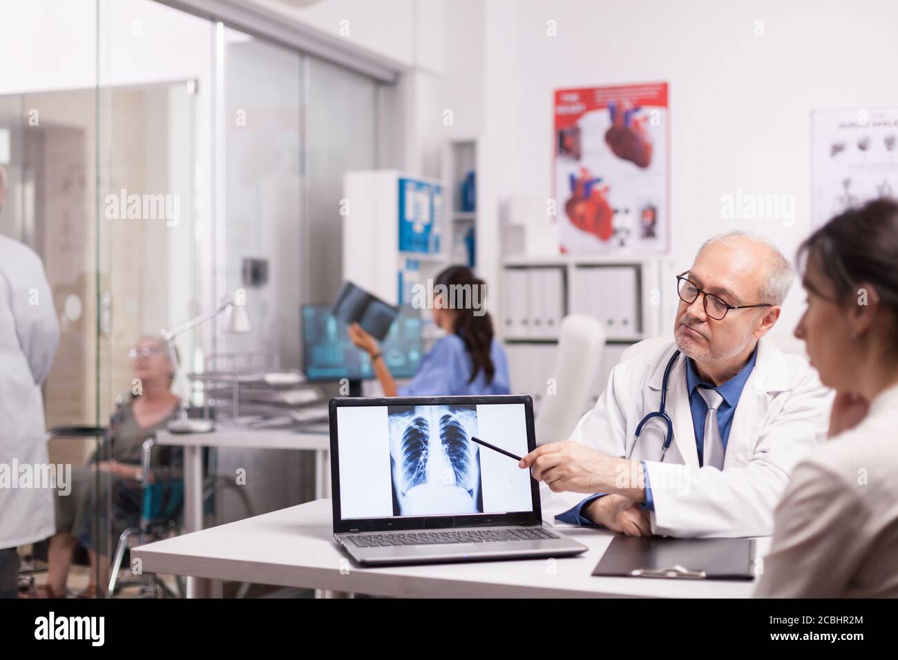 Medico senior che indica la radiografia dei polmoni sani su un computer portatile in ospedale durante la visita medica del paziente. Giovane medico che spiega la diagnosi per disabilitare la donna matura in sedia a rotelle. Foto Stock