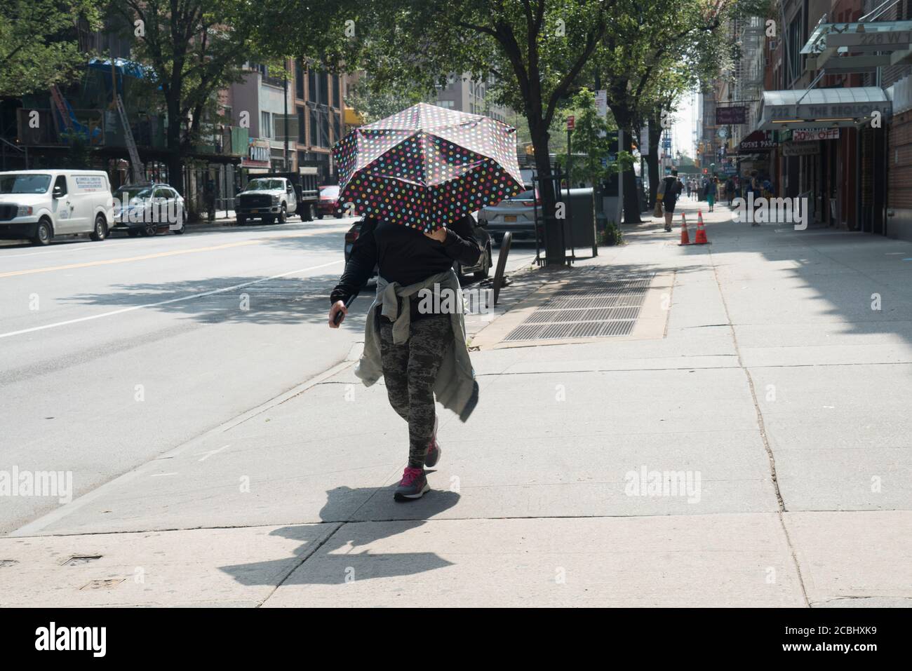 Una donna camminò lungo la quattordicesima strada ovest di Manhattan tenendo un ombrello per proteggersi dal sole. Covid-19 aveva lasciato la strada un tempo trafficata quasi vuota. Foto Stock