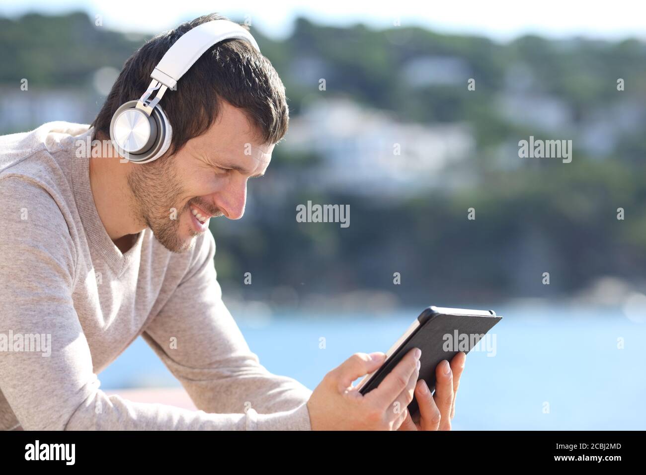 Buon uomo che ascolta la musica con le cuffie wireless su un tablet seduto sulla spiaggia Foto Stock
