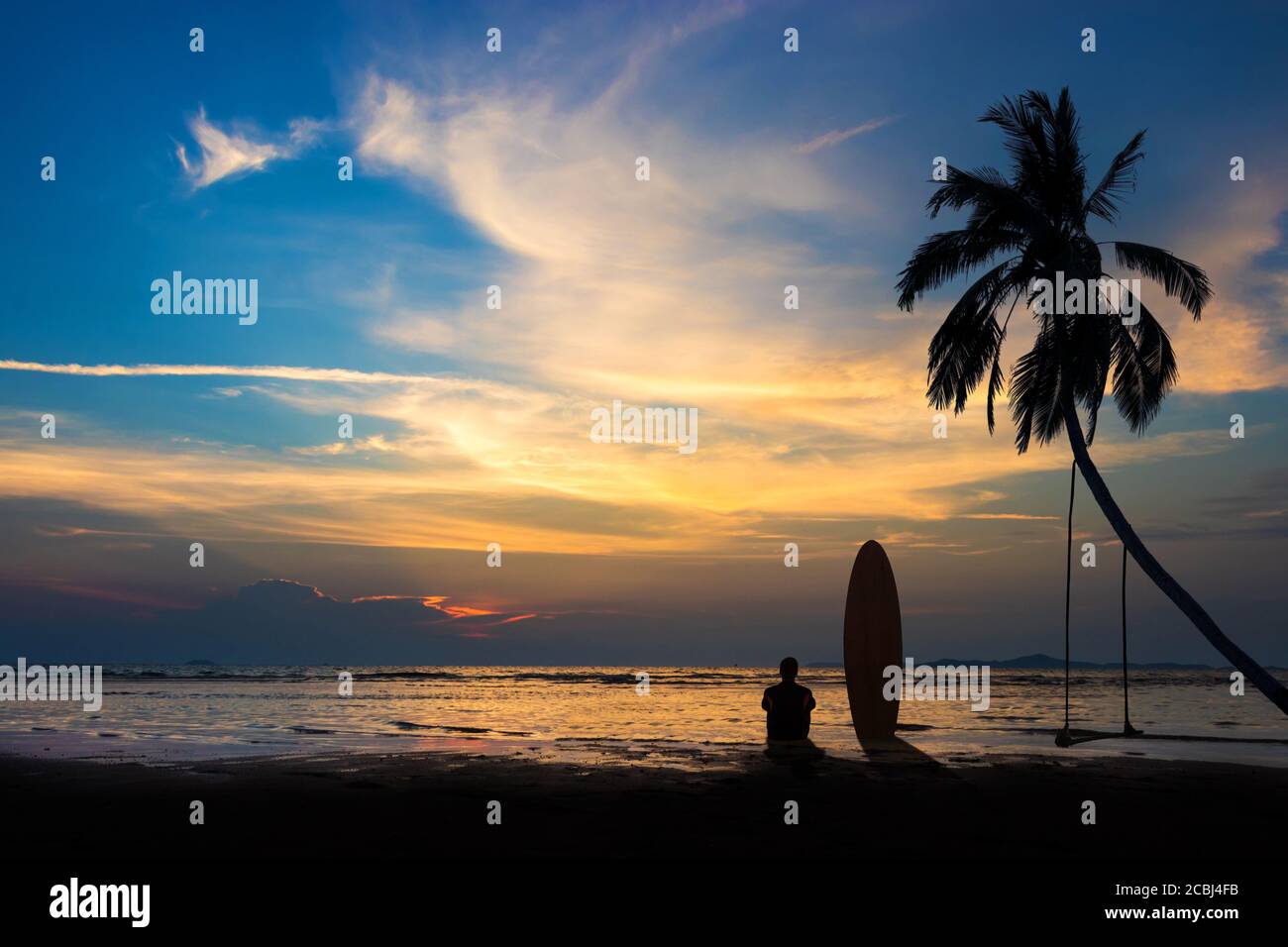 Silhouette di surf uomo sedersi con una tavola da surf sulla spiaggia. Scena di surf alla spiaggia del tramonto con cielo colorato. Sport acquatici all'aperto avventura stile di vita. Foto Stock
